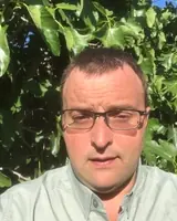 Man with glasses outdoors among green leaves, sunlight on face.