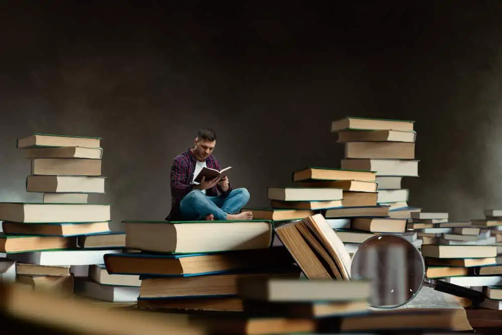 Young man sitting on large stack of books, reading intently in a dark, moody setting.
