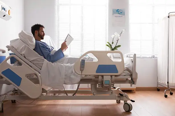 Patient reading a book in hospital bed with medical equipment nearby.