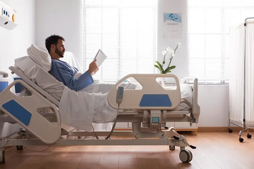 Patient reading a book in hospital bed with medical equipment nearby.