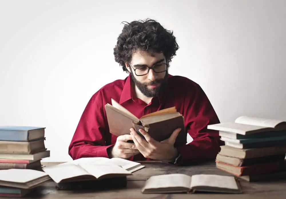 Man reading a book surrounded by numerous open and closed books on a desk.