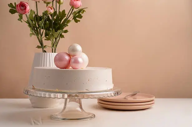 Pastel cake decorated with pink and white spheres, displayed on a glass cake stand with pink plates.