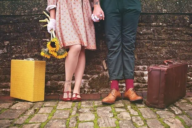 Vintage couple holding hands with travel luggage and flowers.