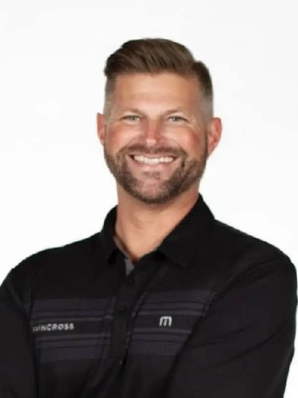 Smiling man with a beard and short hair, wearing a black collared shirt, posing confidently against.