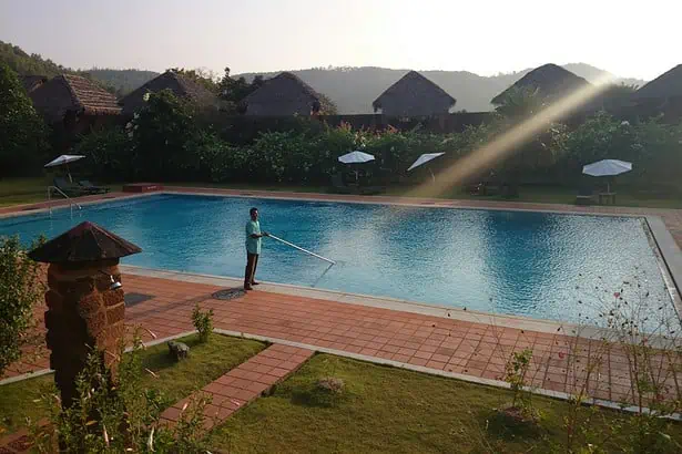 A person cleaning a large outdoor swimming pool surrounded by lush greenery and traditional huts in.