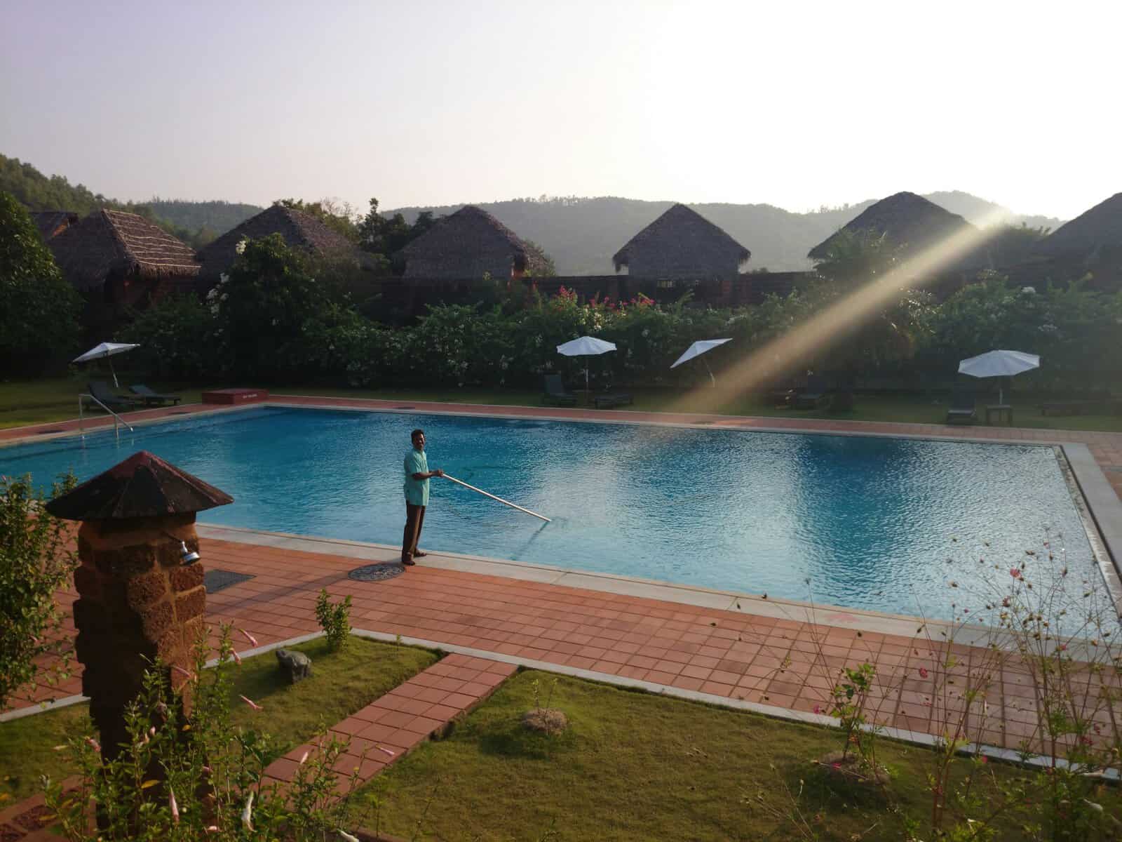 A person cleaning a large outdoor swimming pool surrounded by lush greenery and traditional huts in.