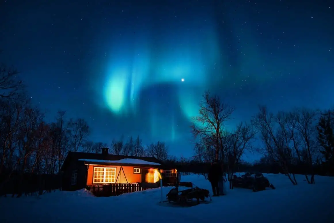 Aurora borealis lights up the night sky above a snow-covered cabin in a winter setting.