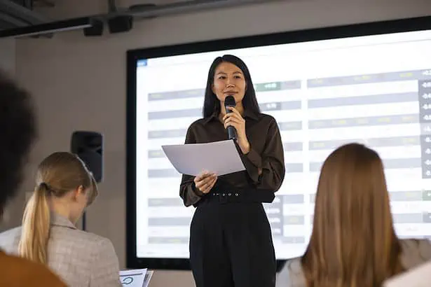 Businesswoman speaking into a microphone during a presentation with a large digital screen behind he.