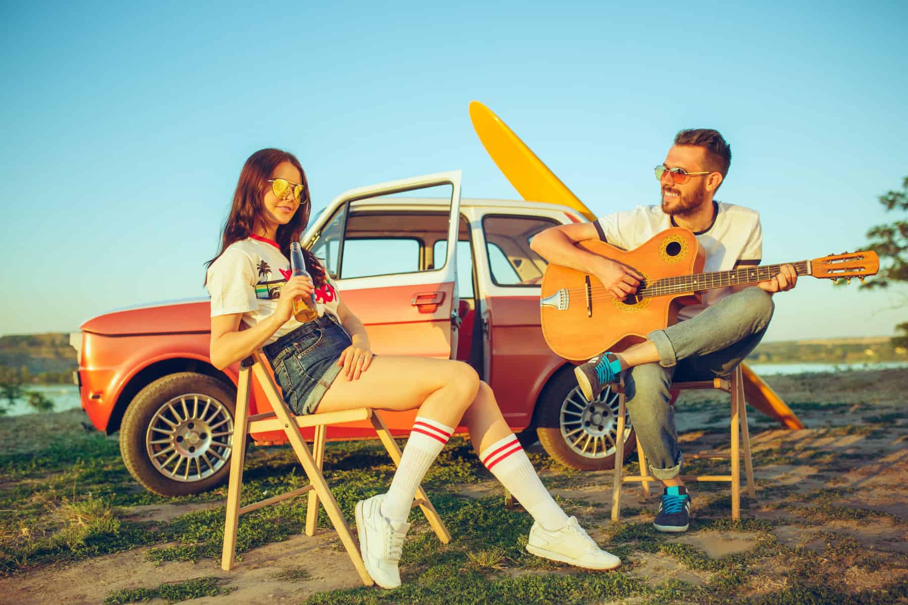 Young woman and man enjoying music by the vintage car with surfboard on a sunny beach day.