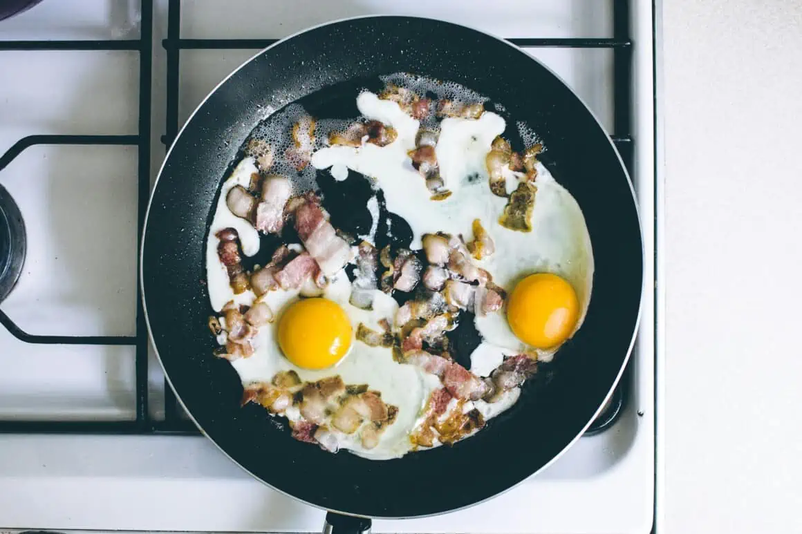 Bacon and eggs frying in a black skillet on a stovetop.