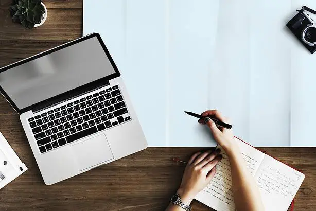 Work desk with open laptop, notebook, pen, and camera on wooden surface.
