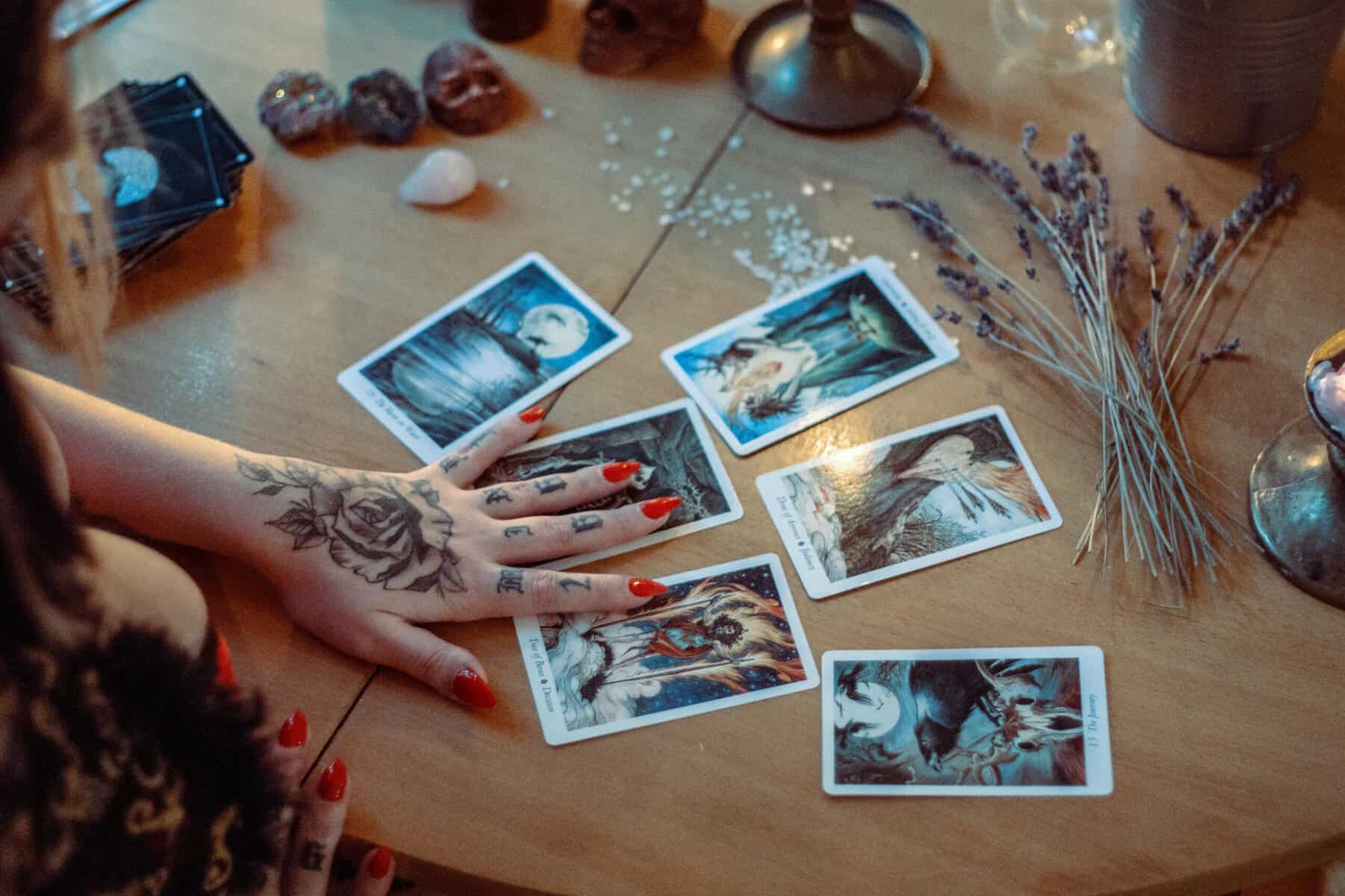 Home Icy 42 An individual with red nails and tattoos performs a tarot card reading on a wooden table, surrounded.