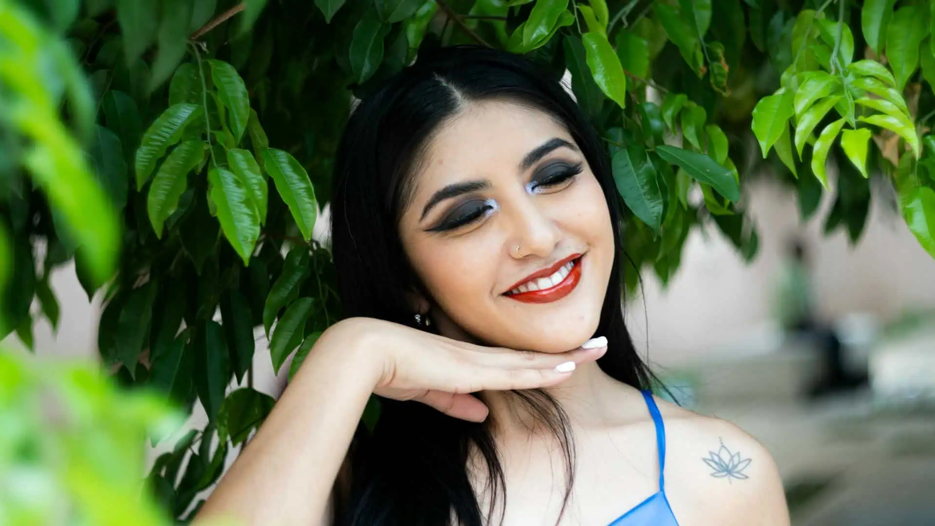 Smiling woman with dark hair and makeup, surrounded by lush green leaves, showcasing natural beauty.