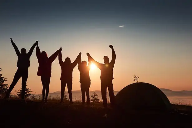 Sunset camping scene with friends holding hands and raising arms near a tent by the lake.