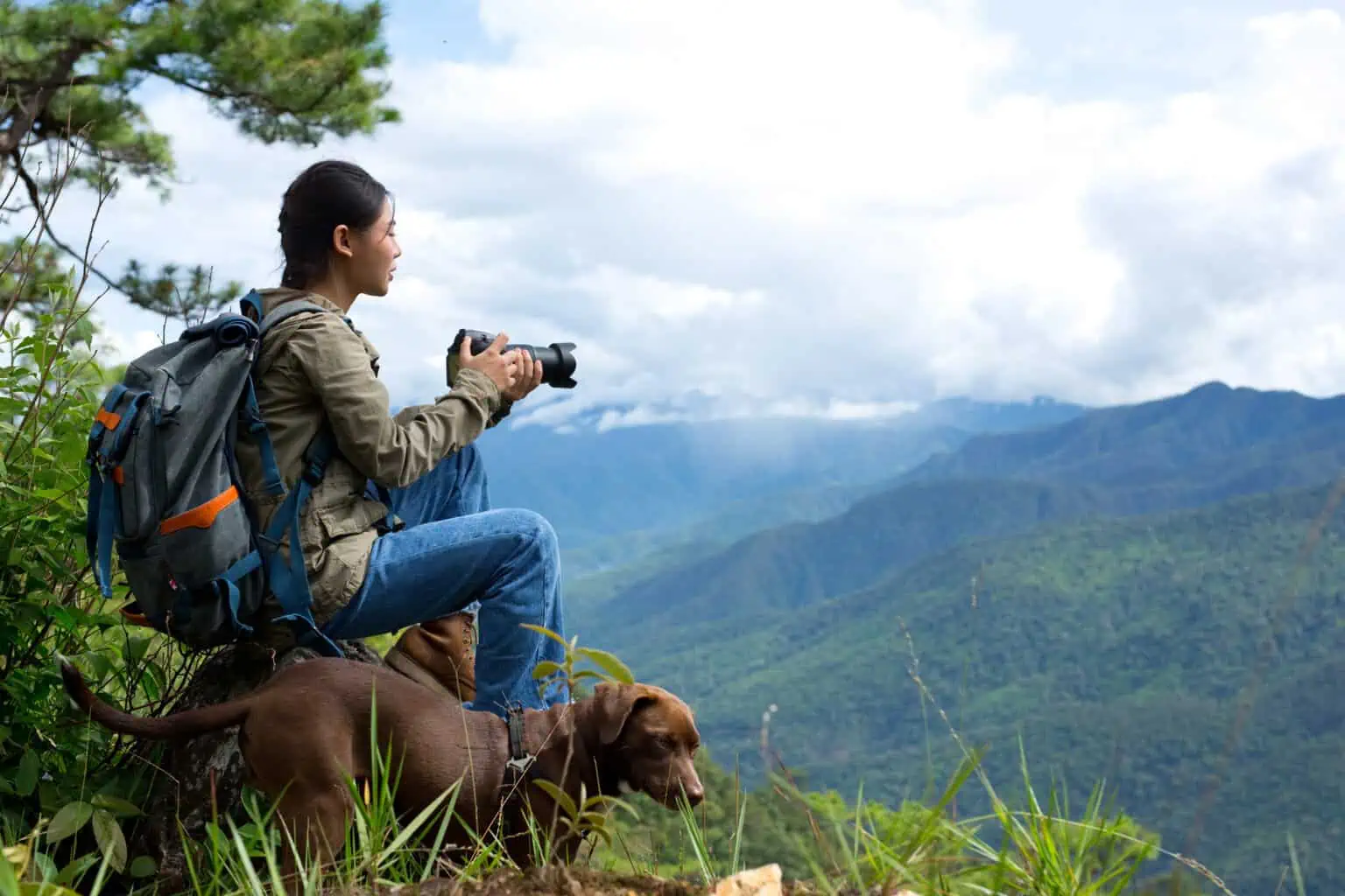 Hiker with backpack taking photos of scenic mountain view with her dog sitting nearby.