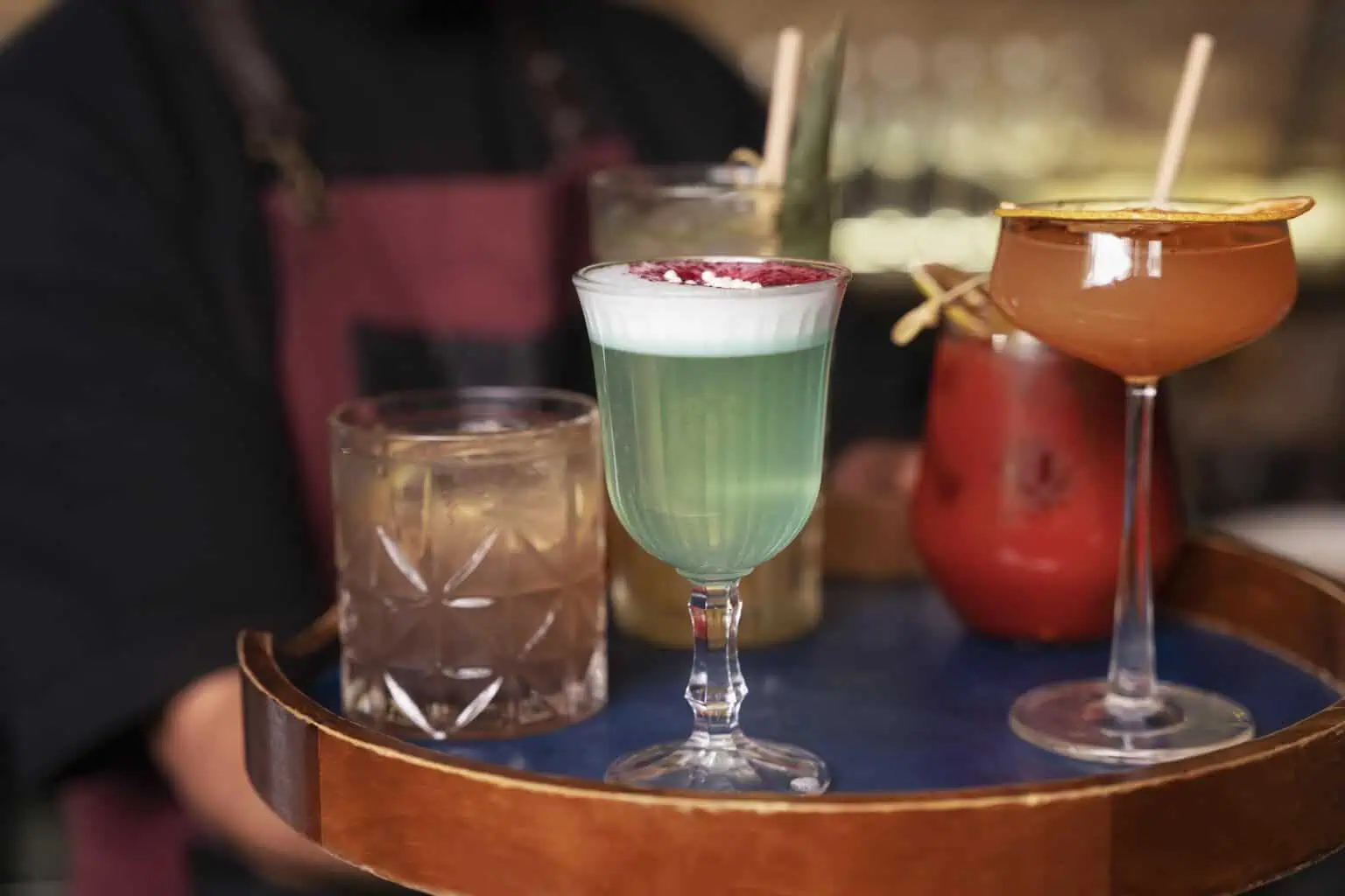 Vibrant cocktails with colorful layers and garnishes on a tray at a lively bar.