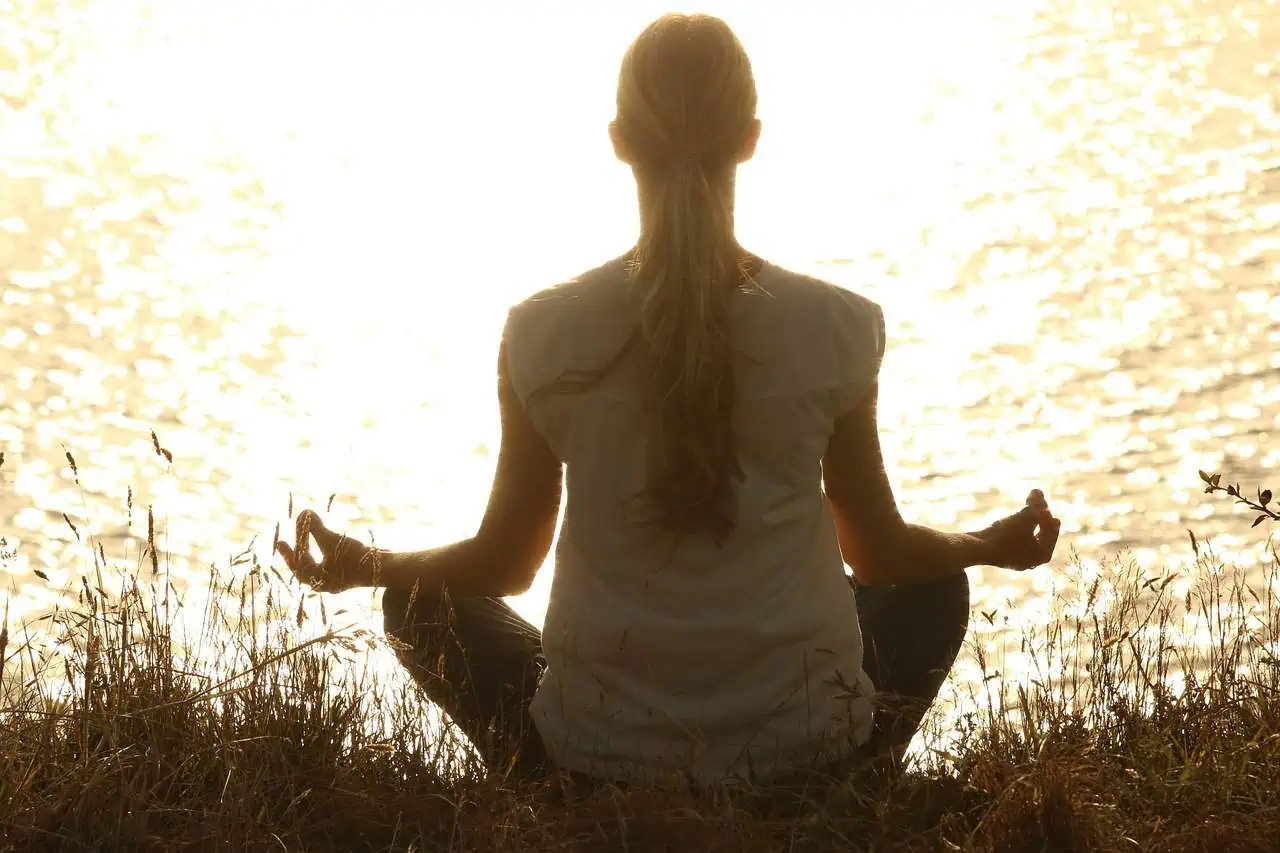 Meditating woman sitting cross-legged by the water during sunset, practicing mindfulness and relaxat.
