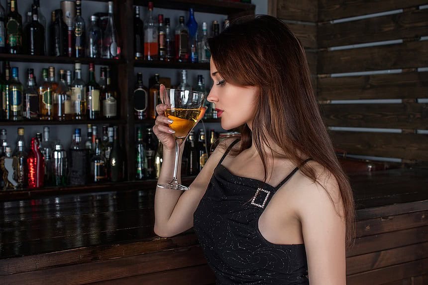 6 Best Luxury Drug and Alcohol Detox Centers in Los Angeles (2026) 1 A woman enjoying a cocktail at a bar with shelves of liquor bottles in the background.
