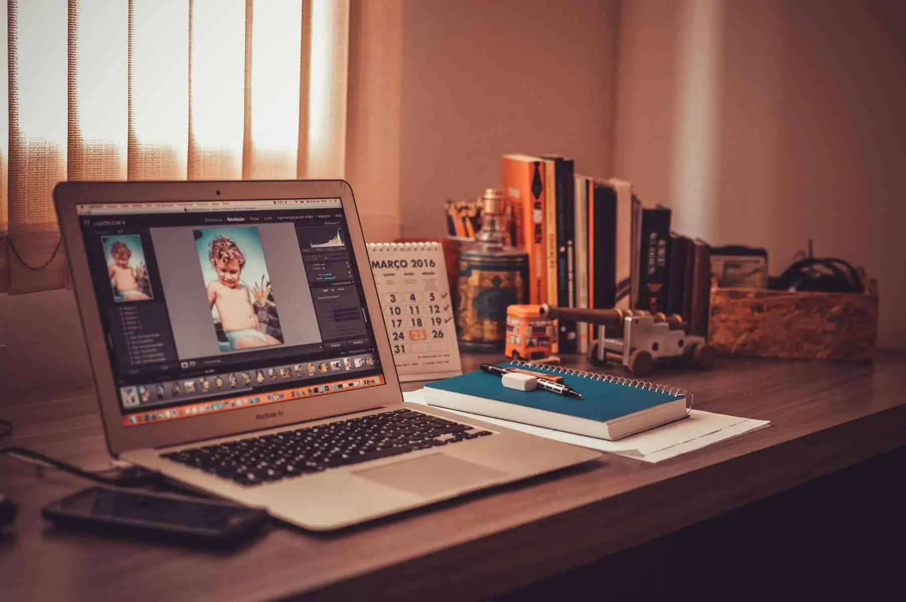 Laptop workspace with photo editing tools and books on a wooden desk.
