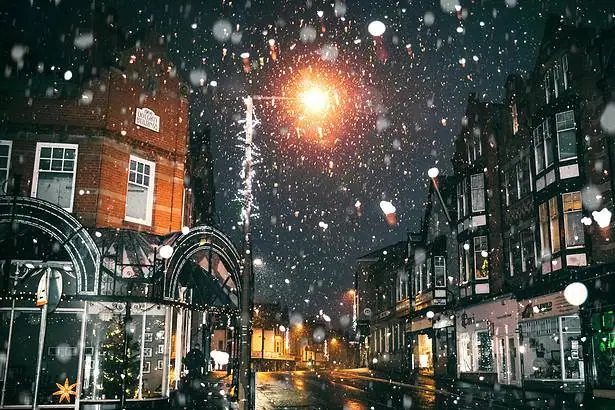 Snow falling on a city street illuminated by streetlights at night.
