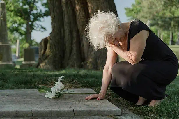 Elderly woman mourning at a gravesite with flowers in a peaceful cemetery.