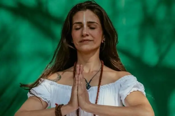 Woman meditating outdoors with hands in prayer position, surrounded by lush green trees.
