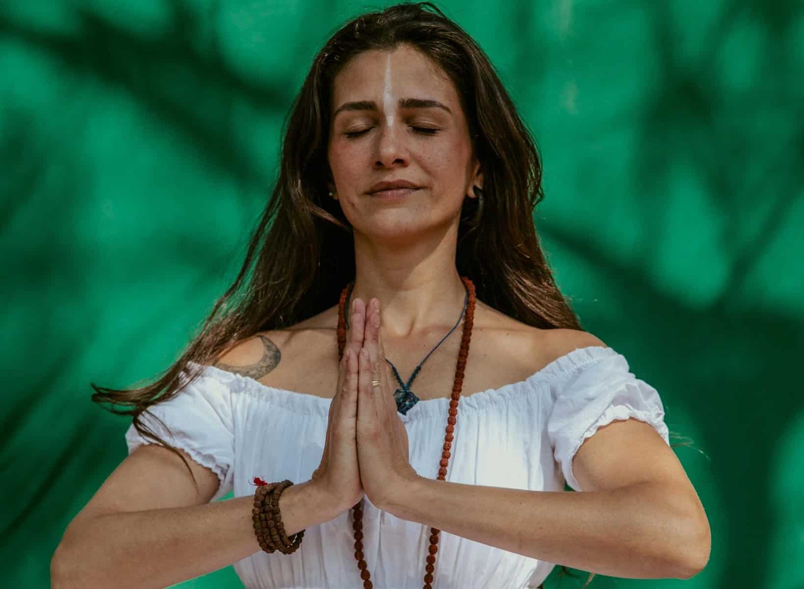 Woman meditating outdoors with hands in prayer position, surrounded by lush green trees.