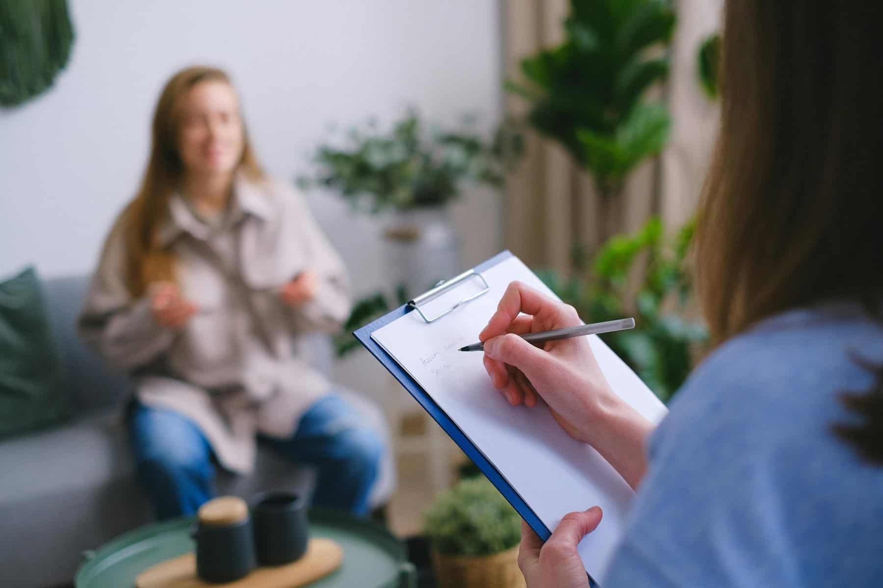 Home Icy 48 Therapist taking notes during a therapy session with a young woman in a cozy, well-lit room.
