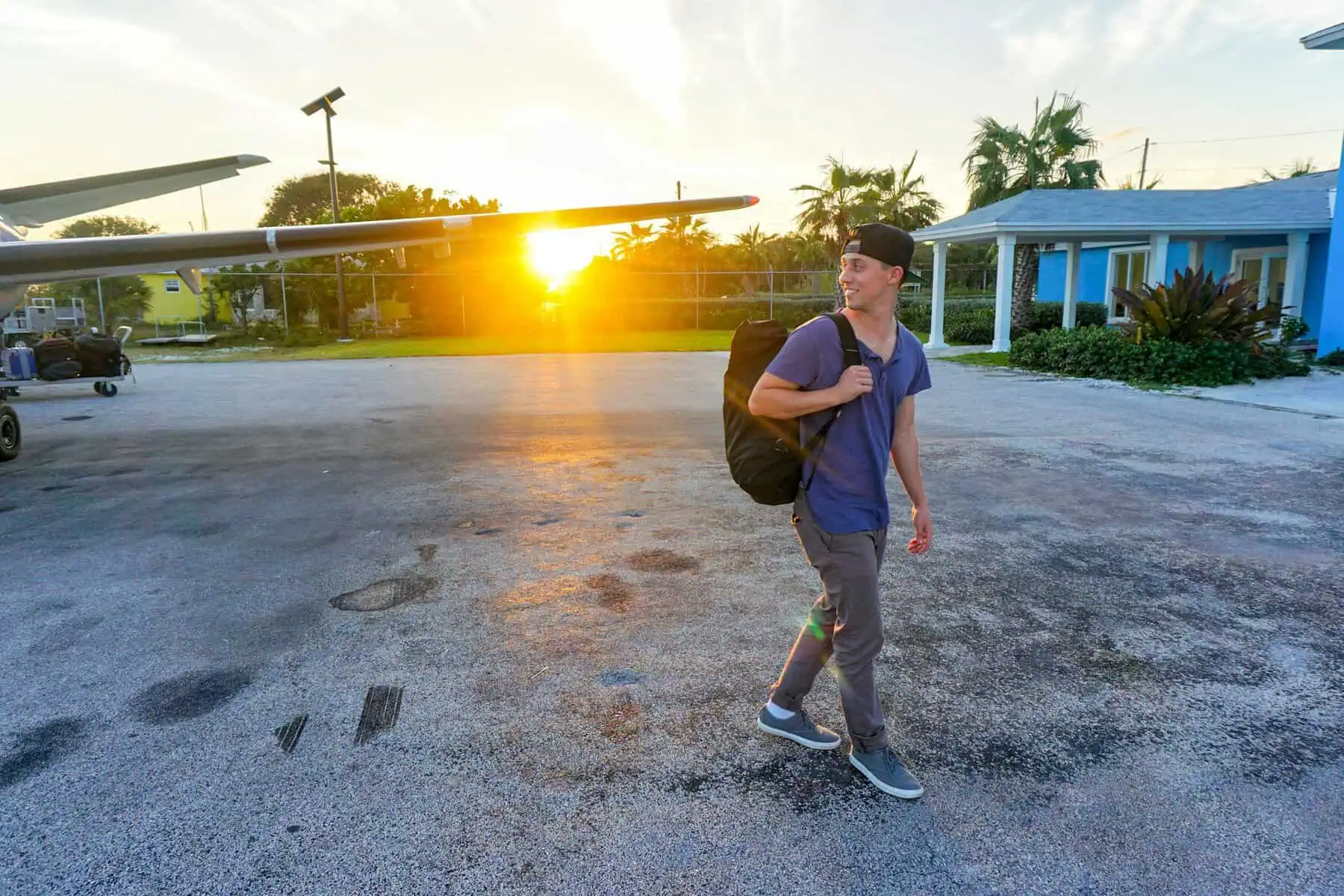 Young man with backpack walking on tarmac at small airport during sunset.