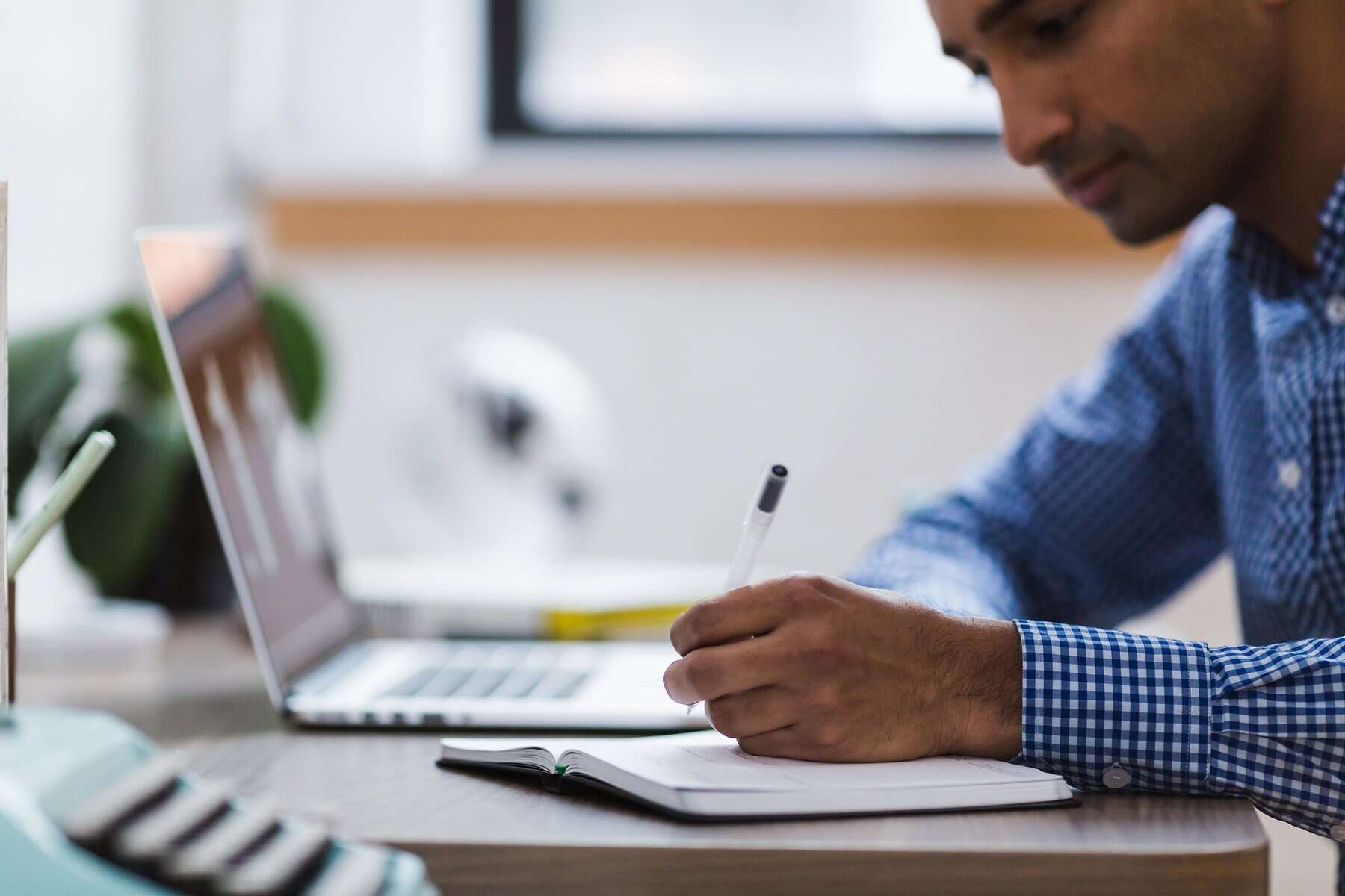 Person writing with a pen on a notebook at a desk with laptop and plant.