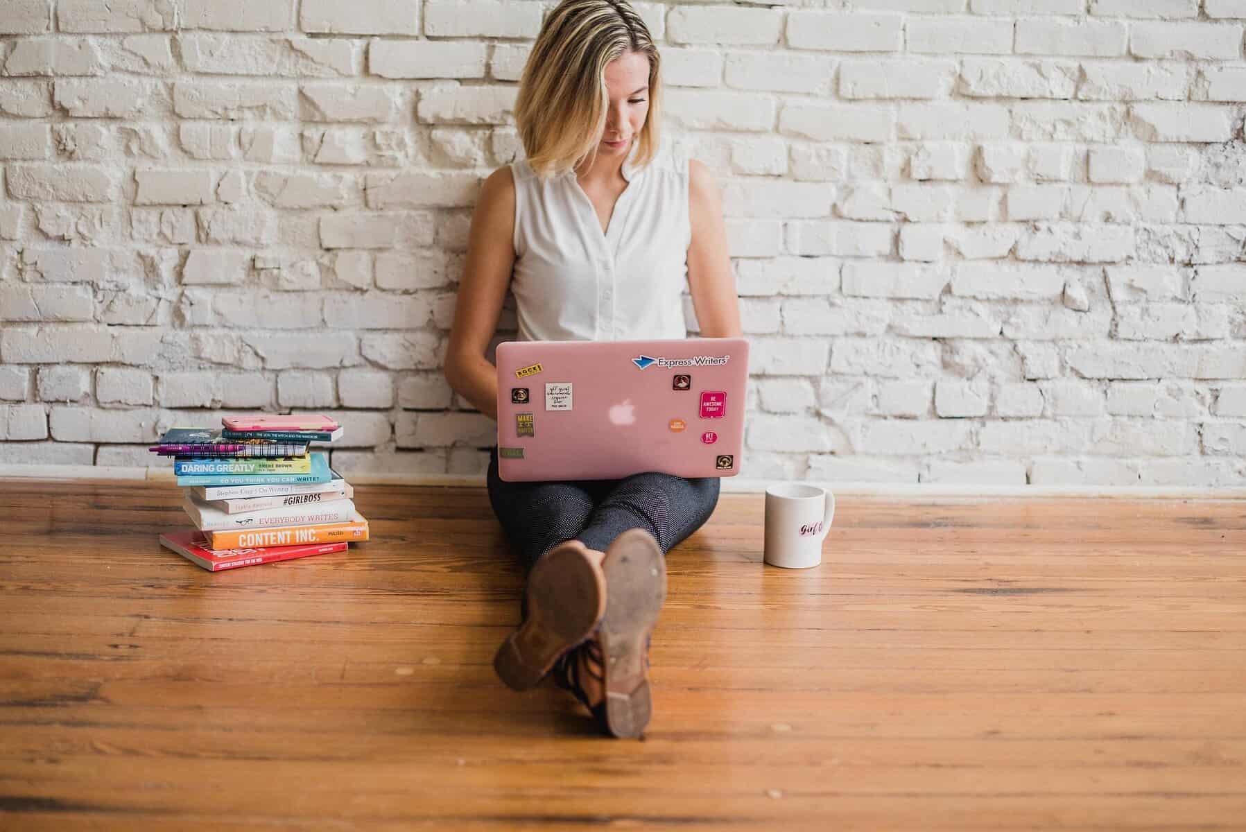 Home Icy 39 Alt text: Woman sitting on wooden floor with laptop, books, and mug against white brick wall.