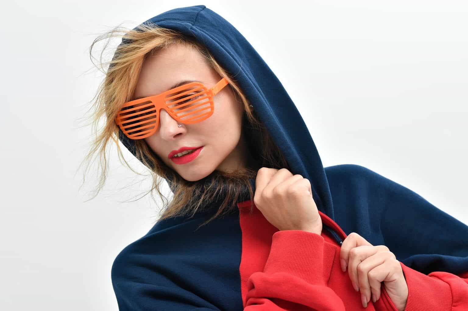 Stylish young woman wearing a navy hoodie and trendy orange shutter sunglasses, vibrant makeup, against plain background.