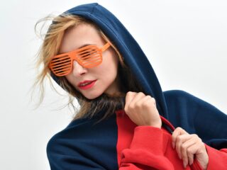 Stylish young woman wearing a navy hoodie and trendy orange shutter sunglasses, vibrant makeup, against plain background.