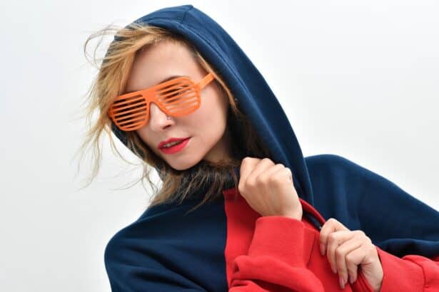 Stylish young woman wearing a navy hoodie and trendy orange shutter sunglasses, vibrant makeup, against plain background.