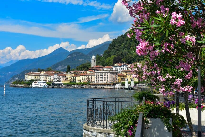 Stunning lakeside view featuring vibrant pink flowers, charming Italian architecture, and majestic mountains under a bright blue sky.